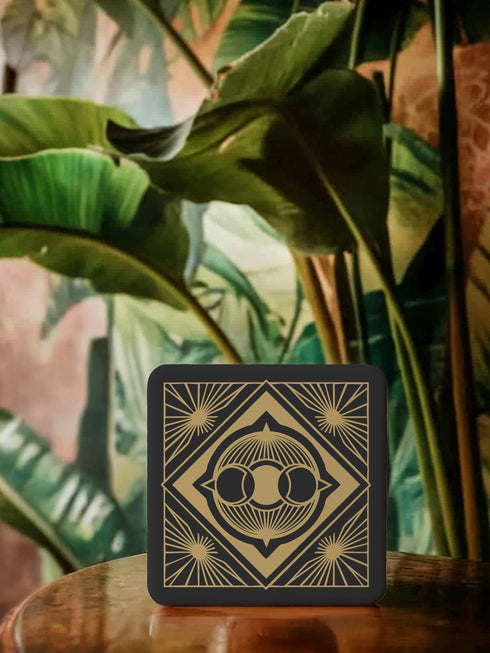 A black and gold ceramic coaster featuring a triple moon design framed by art deco geometric details and celestial sunburst accents.